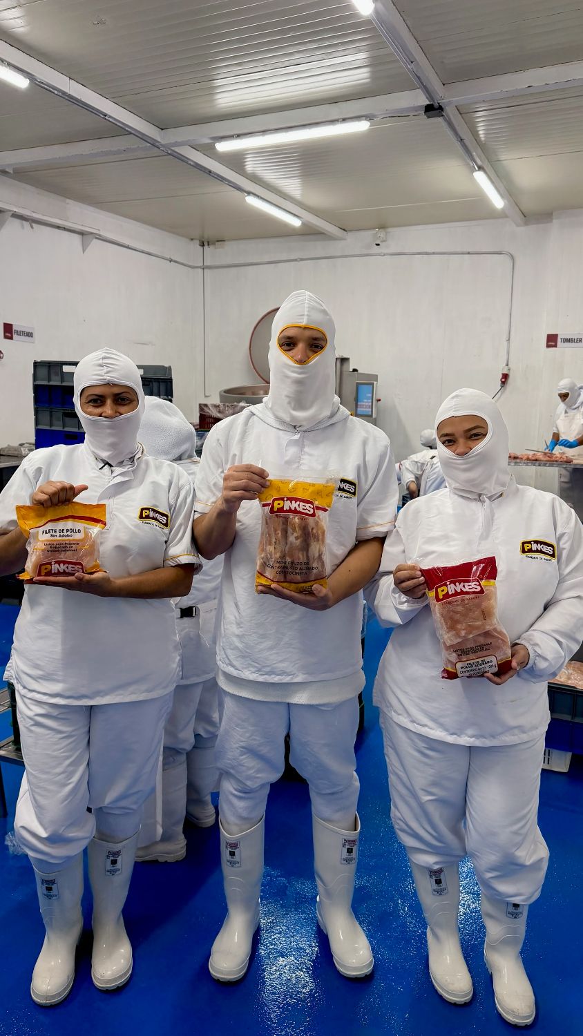 Tres integrantes del equipo Pinkes con uniforme completo sostienen las bolsas de filete de pollo sin adobo, mini chuzo de contramuslo con tocineta y filete de pollo adobado en la planta