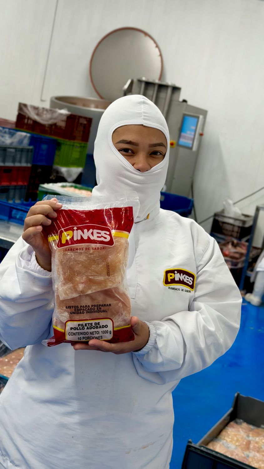 Operaria de Pinkes con uniforme completo sostiene una bolsa de filete de pollo adobado frente al tómbler de la planta
